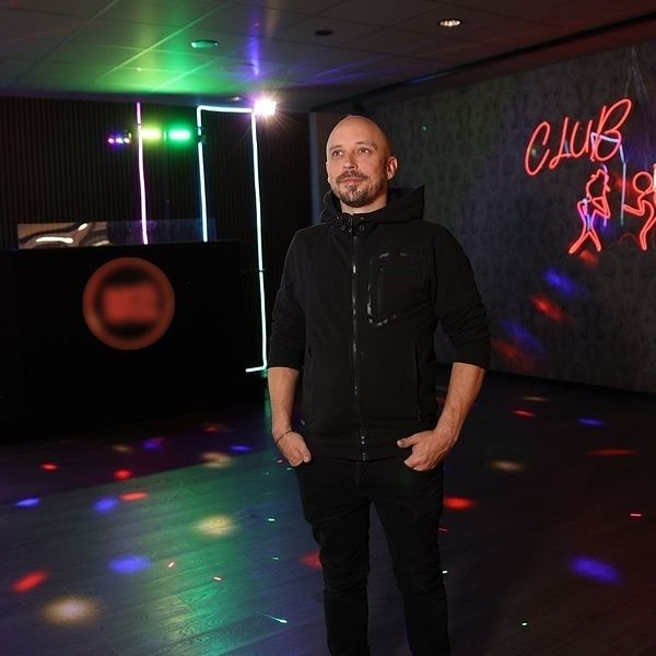 En person står i ett mörkt rum med färgglad belysning, inklusive blinkande ljus och neonskyltar. Rummet har en DJ-bås med en rund logotyp synlig. På väggen finns en neonskylt med illustrationer av dansande figurer. Atmosfären ger en känsla av nattliv och fest.