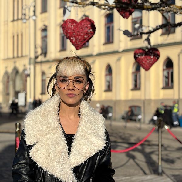 En kvinna med ljus pälsjacka står framför en byggnad och ett träd dekorerat med röda hjärtan hänger från grenarna.