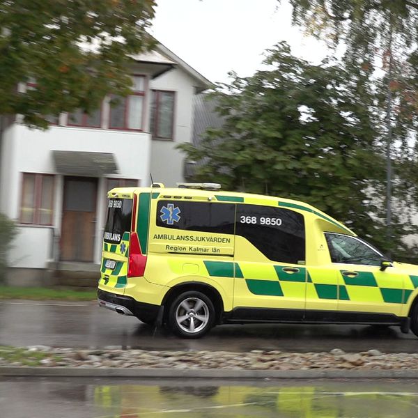 Ambulans på regnvåt asfalt. Träd och ett vitt hus i bakgrunden.