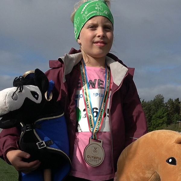 Bilden visar flicka med medalj runt halsen och två käpphästar