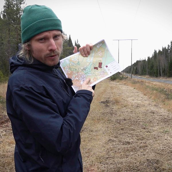 SVTs reporter står längs ett motionsspår omgiven av skog, hållande en karta och pekar på den. I bakgrunden syns kraftledningar som sträcker sig längs vägen.