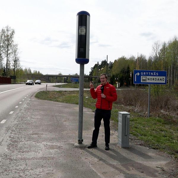 Reporter står med röd jacka och pekar på en fartkamera intill en väg.