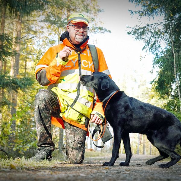 En person i orange och gul arbetsjacka står på knä i en skogsmiljö och interagerar med en svart hund som står bredvid. Området är omgivet av träd och ger intryck av att vara en friluftsmiljö, där personen verkar ta en paus för att umgås med hunden.