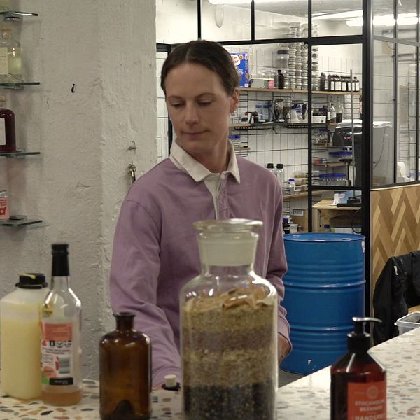 Person står bakom en bardisk med olika flaskor och burkar framför sig i en modern lokal.