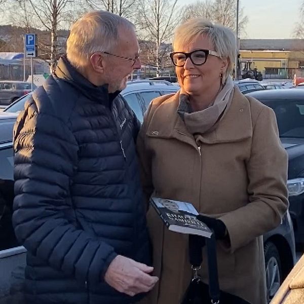 Två personer står nära varandra på en parkeringsplats, omgivna av bilar. En av dem håller en bok, och det är vinter med snö på marken. I bakgrunden syns träd och en byggnad.