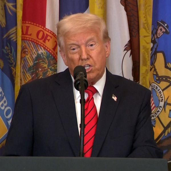 Person i kostym står vid podium framför USA-delstatsflaggor.