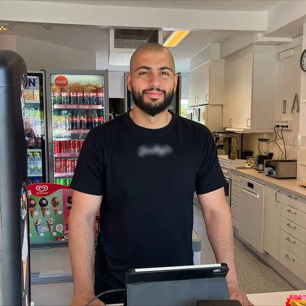 Person bakom kassan i en kiosk med dryckeskylar och glassmaskin i bakgrunden.