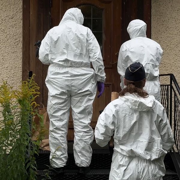 Tre personer i skyddsdräkter står på en trappa framför en dörr. Personerna är vända bort från kameran, med en av dem som bär handskar och en annan med en mössa. Området runt trappen har grön vegetation.