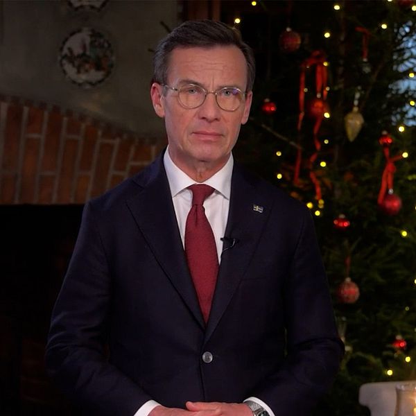 Ulf Kristersson står framför en tänd brasa, medan en julgran lyses upp i bakgrunden. Stämningen är varm och julig.
