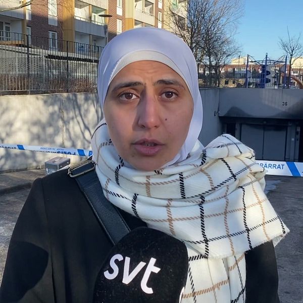 En person iförd en svart jacka och vit- och svartmönstrad sjal står framför ett avspärrat område med polistejp. Bakgrunden visar en porten till ett underjordisk garage, bostadshus och en klarblå himmel. En mikrofon med SVT:s logotyp hålls av personen.