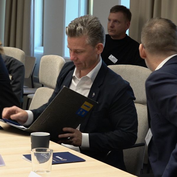 En grupp personer sitter runt ett konferensbord och diskuterar, med mappar och kaffe framför sig. Namnskyltar är placerade på bordet.