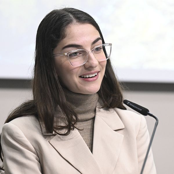 Kvinna i ljus kavaj står vid en mikrofon framför en presentationsduk.