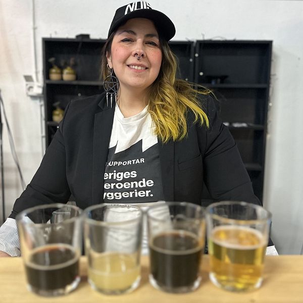 En kvinna med en t-shirt med texten ”Sveriges oberoende bryggerier” och svart kavaj och keps står bakom en bardisk.