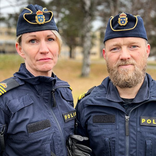Två poliser poserar tillsammans utomhus, klädda i mörkblå uniformer med svenska polisens insignier. Bakgrunden visar en park med träd och en byggnad i fjärran, vilket antyder en lugn miljö.