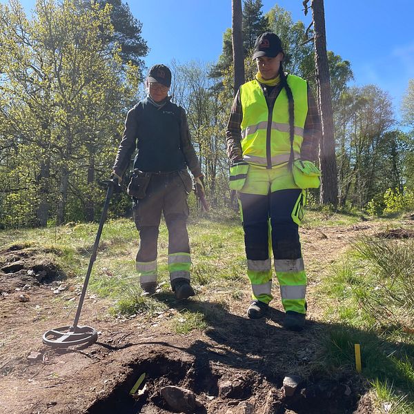 Två arkeologer söker med metalldetektor i en solig skogsglänta i Värnamo.