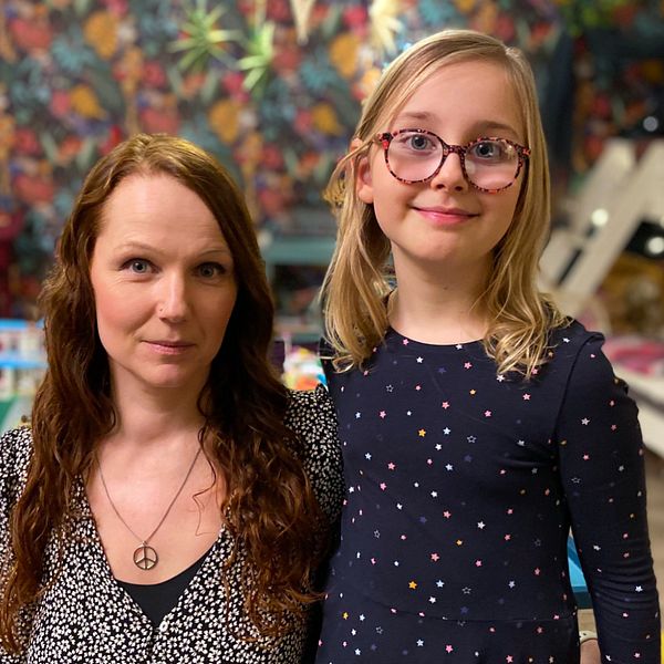 En kvinna och en liten flicka står tillsammans i ett färgglatt rum med mönstrad tapet och leksaker på golvet. Rummet har en familjär atmosfär med en säng och en stol i bakgrunden.