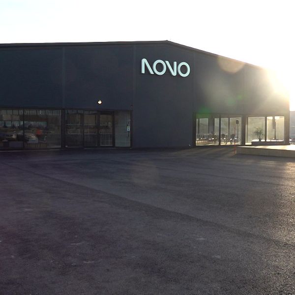 Industrihall med skylten NOVO, tom parkeringsyta och motljus från solnedgång.