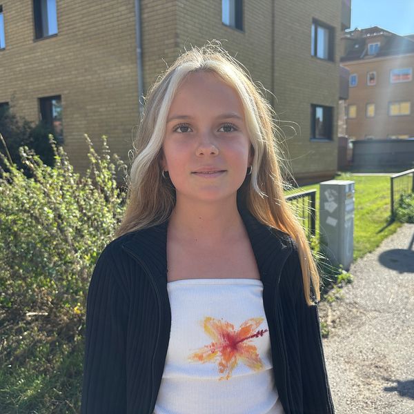 En person med långt blont hår står på en gata omgiven av gröna buskar och byggnader. Personen bär en mörk kavaj över en ljus top med en blommig design. I bakgrunden syns en trottoar och en parkerad elscooter. Soligt väder skapar en ljus och vänlig atmosfär.
