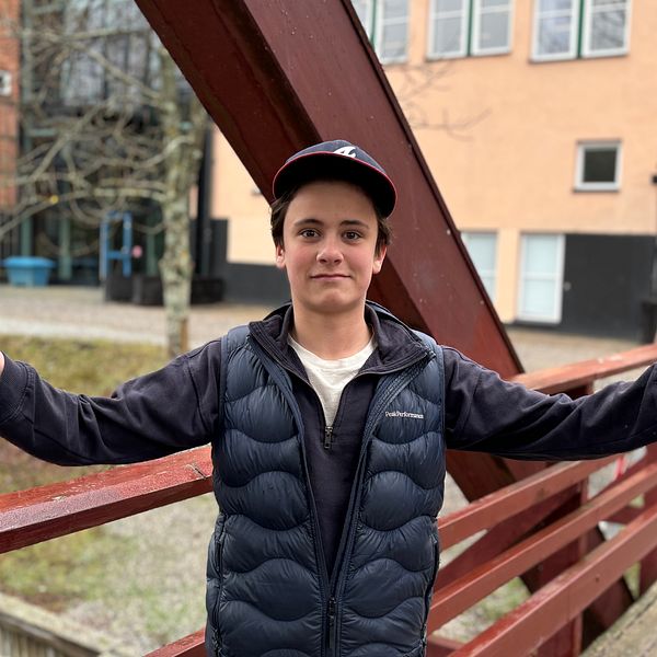 Person med keps står på bro med utsträckta armar, byggnader syns i bakgrunden.