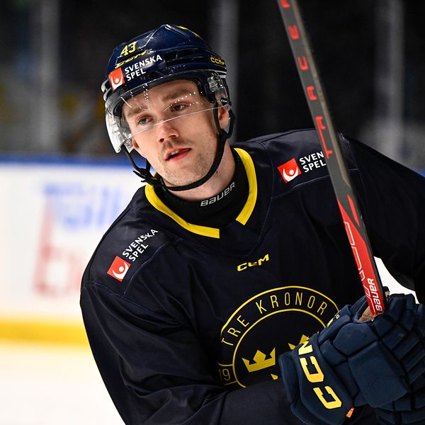 En ishockeyspelare från Tre Kronor i mörkblå utrustning och hjälm, håller en röd ishockeyklubba vid sidan av isen med reklamskyltar i bakgrunden.