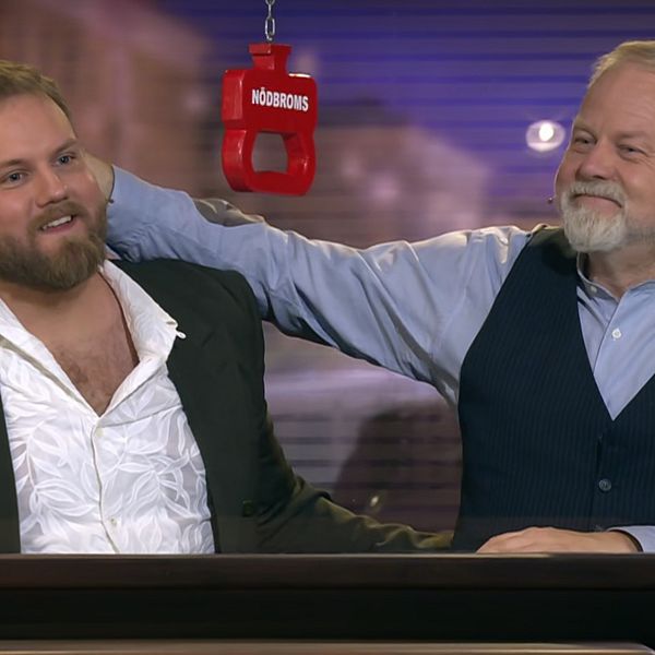 Två män sitter vid ett bord i en studiomiljö med en mikrofon hängande mellan dem. En av männen har sin arm över den andres axlar.