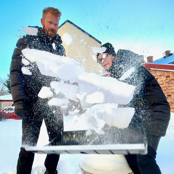 Två personer står i snön, den ena skyfflar snö mot kameran.