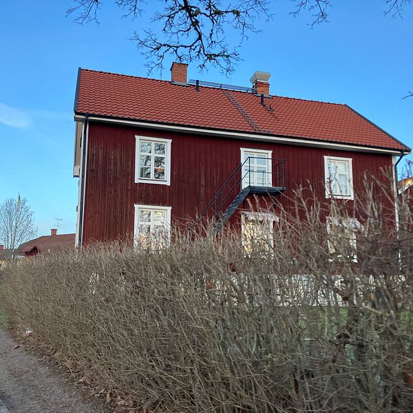 Ett rödmålat hus med vitmålade fönster och en balkong, omgiven av en tät häck och beläget vid en grusväg. Himlen är klar blå och det finns träd i bakgrunden.