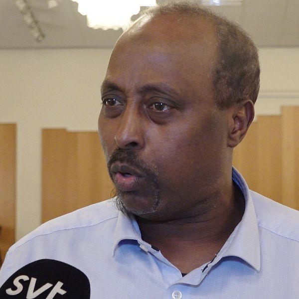 En person står i ett rum med träpaneler på väggarna. I förgrunden hålls en mikrofon märkt med ”SVT”, vilket tyder på en intervjusituation i en studiomiljö eller ett offentligt uttalande.