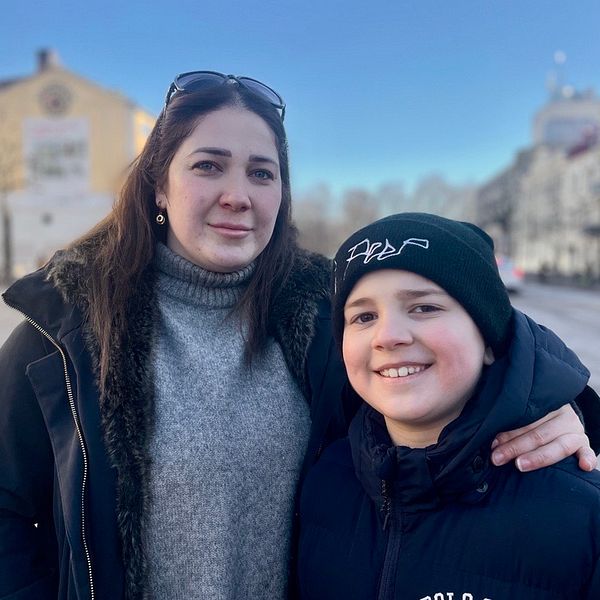 Två personer står tillsammans på en trottoar i en stadsmiljö. Den ena personen bär solglasögon och har en arm runt den andra personen, som är klädd i en mörk jacka och mössa. Bakgrunden visar en gata med byggnader och träd under en klarblå himmel.