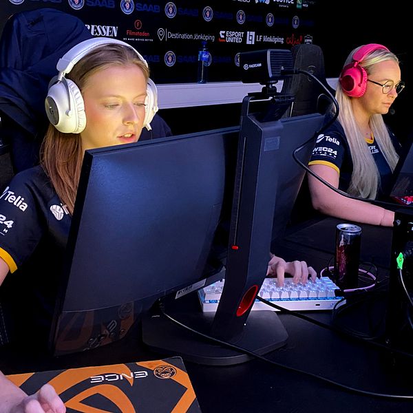 Två kvinnliga gamers sitter vid sina datorer under en e-sporttävling. Landslagspelarna Emma Mattsson och Linnéa Claesson. Den första spelaren har på sig hörlurar och är fokuserad på skärmen, medan den andra också bär hörlurar och använder ett tangentbord. Båda spelar i en mörk miljö med lagets logotyper synliga i bakgrunden.