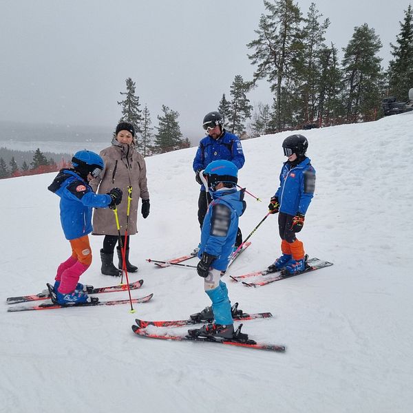 En grupp barn i skidkläder står på snön vid en skidbacke. Två av barnen är upptagna med att justera sina skidor medan den vuxna och andra barn tittar på. Det snöar lätt och det syns träd i bakgrunden.
