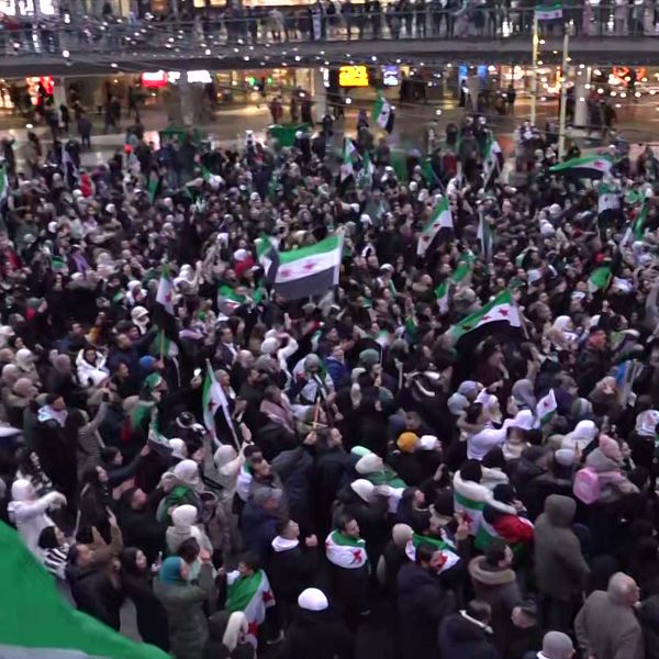 Stort folkmassa som håller upp syriska flaggor under en demonstration. Människorna är samlade på en offentlig plats, klädda i varierande kläder, och det är en känsla av gemenskap och engagemang. Bakgrunden visar en livlig stadsbild med belysning och byggnader.