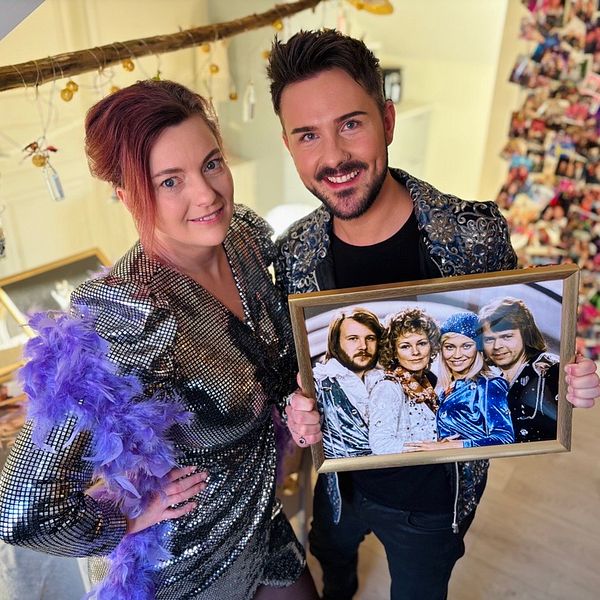 Två personer klädda i glittriga kläder håller i en inramad fotografi med Abba. De står i ett utrymme dekorerat med en vajer fylld med bilder och färgglada klädnypor i bakgrunden.