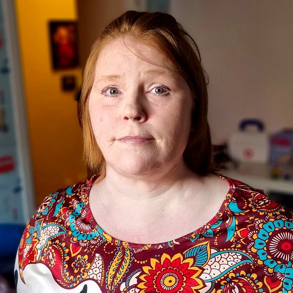 Cecilia, förälder som har svårt att få ekonomin att gå ihop, står i ett rum klädd i en färgglad blommig blus. I bakgrunden syns väggar dekorerade med bilder av bilar. På en hylla i bakgrunden finns förvaringslådor och leksaker.