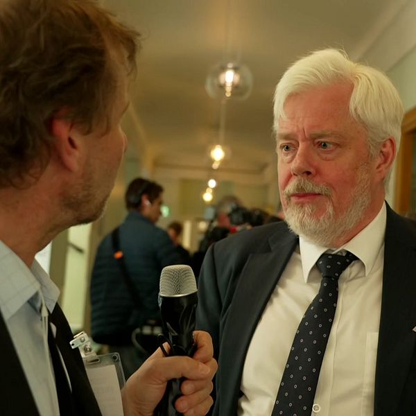 Reporter intervjuar Niklas Ekvall i kostym i en korridor, flera personer syns i bakgrunden.