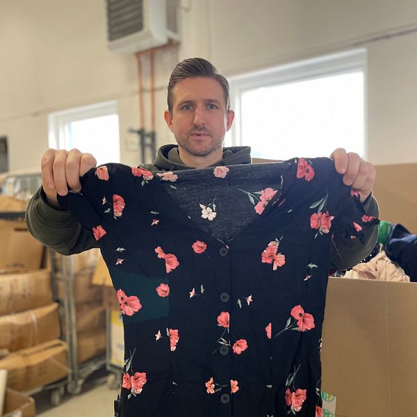 En person håller upp en svart klänning med röda blommor i vad som verkar vara ett lager- eller secondhand-miljö, omgivet av stora kartonger och lagerhyllor.