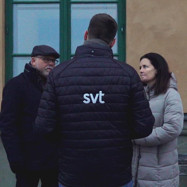En reporter från SVT intervjuar två personer utomhus vid en byggnad med grön fönsterlucka. Reportern bär en svart jacka med SVT-logotyp på ryggen.
