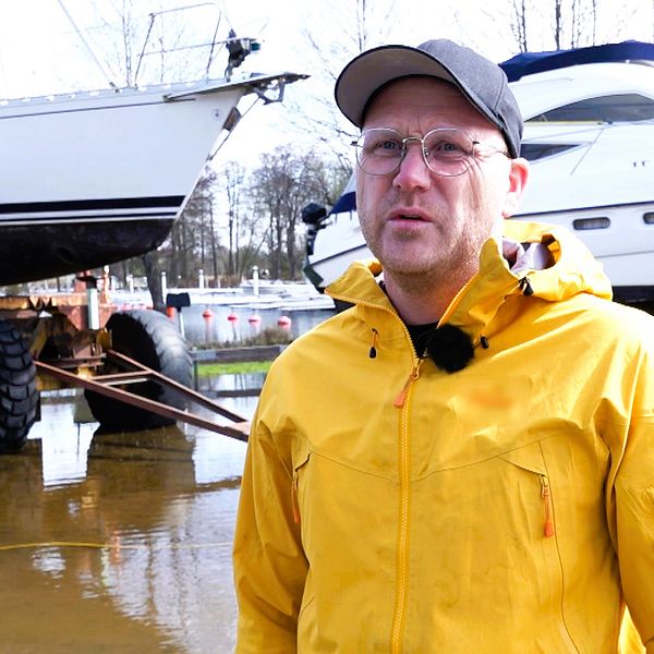 Man i gul jacka med svart keps. Står på land framför båt. I bakgrunden syns översvämmat mark.