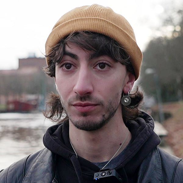 Louka Bertolino har en senapsgul mössa på sig och örhänge. Han har mörka ögon och skäggstubb. Han har på sig en svart skinnjacka och en svart hoodie under den.