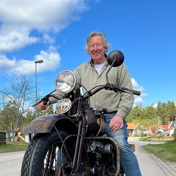 Leende man sitter på en motorcykel på en landsväg med skog och blå himmel i bakgrunden.