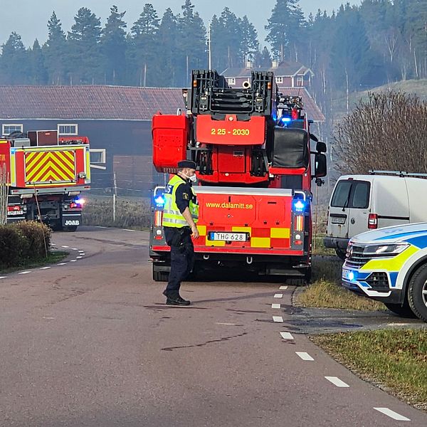 Brandbilar och en polisbil på en väg i Rättvik. En polisman med skydd för ansiktet står på vägen och rök syns i luften.