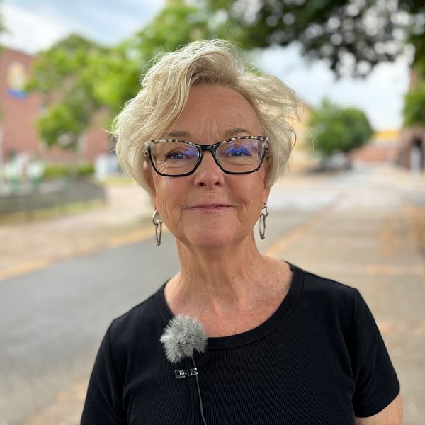 Kvinna med mikrofon står utomhus på en gata med träd och byggnader i bakgrunden.