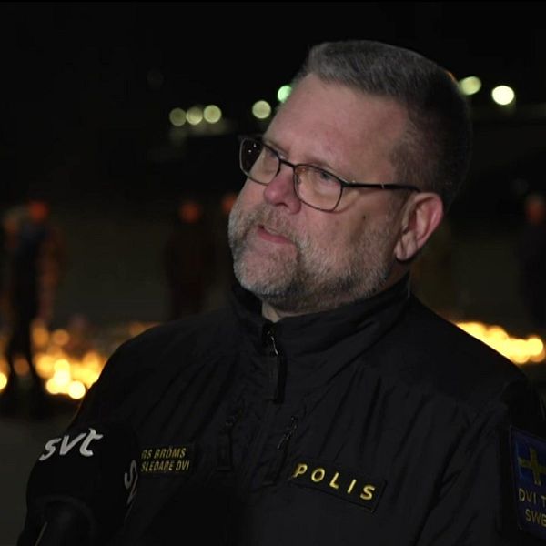 En polis står framför en minnesplats med tända gravljus på en mörk plats, med en mikrofon från SVT i närheten. Olika personer syns i bakgrunden.