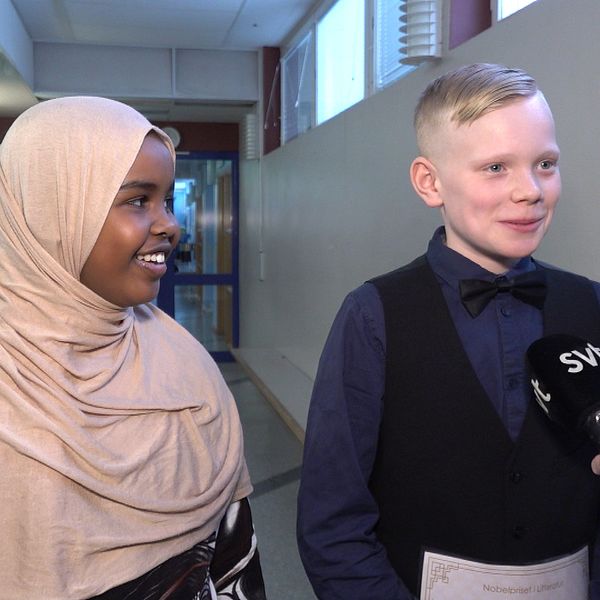 Två barn, en pojke och en flicka, står finklädda och ler i en skolkorridor. Framför sig har de en mikrofon som det står SVT på.