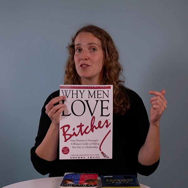 En person håller upp boken ”Why Men Love Bitches” framför sig, med en svart tröja på och en hand gestikulerande i luften. På bordet framför personen ligger två andra böcker synliga. Bakgrunden är ljusblå.