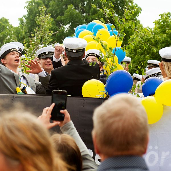 Studenter i vita mössor firar med blågula ballonger och vinkar till publiken