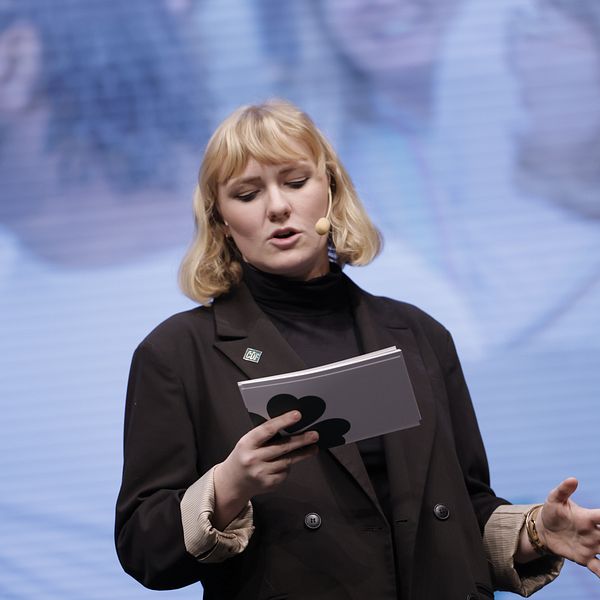 Caroline von Seth talar framför en suddig bakgrund under ett evenemang eller en konferens, med en mikrofon i handen och ett block eller anteckningar i den andra handen.