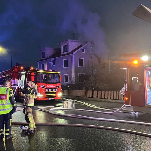 Rök kommer från fönster på andra våningen i tvåvåningshus. Mörkt på platsen med gatlyktor. Räddningstjänst med två bilar och personal på vägen utanför huset.