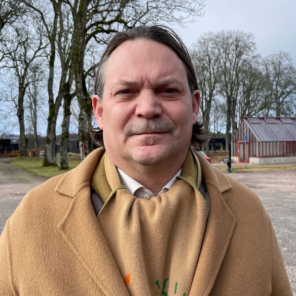 Person i beige kappa står utomhus med träd och byggnader i bakgrunden.