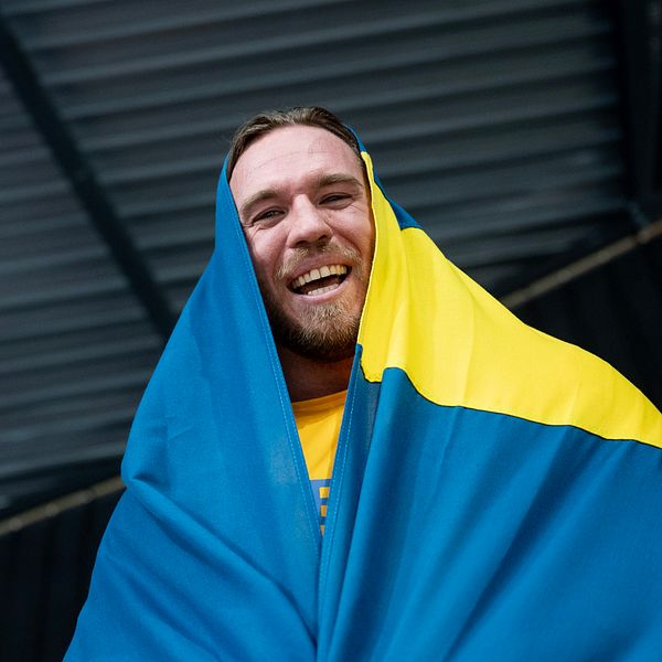 Person inlindad i svensk flagga under ett tak med metallpaneler.